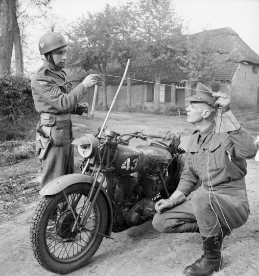 Photo - Corps of Military Police motorcyclists demonstrate how a metal rod fitted to a motorcycle can prevent the rider from being killed by a wire stretched across the road, 25 October 1944. (Image courtesy Imperial War Museum)