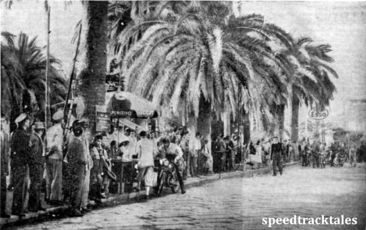 photo - The tropical-like background to the check at Albenga, with J. C. de Wit (125 Eysink), Dutch Trophy team, at the table. Other riders are waiting their turn to check in. ISDT 1948 (Speedtracktales Collection)